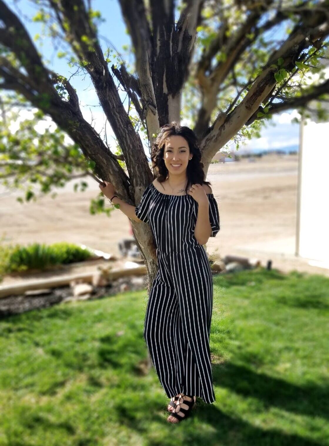 Woman in a black and white vertical stripped jumpsuit standing in front of a tree Smiling