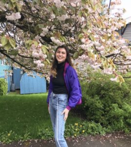A woman wearing a black shirt, purple jacket, and jeans standing in front of a flower tree smiling.